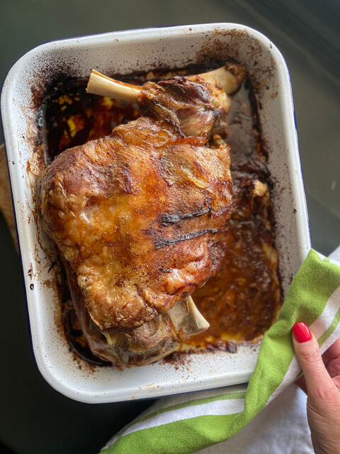 A roast lamb joint with the bone showing in a white enamel dish, held with a green striped cloth