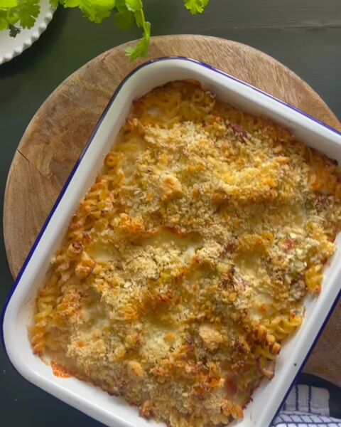 A golden, crumb-topped pasta bake in a white enamel dish with a blue rim, on a wooden board with a blue-checked cloth