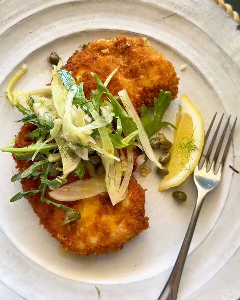 A crumbed chicken escalope on a white plate with a shaved fennel and leaf salad, capers, a lemon wedge and a fork