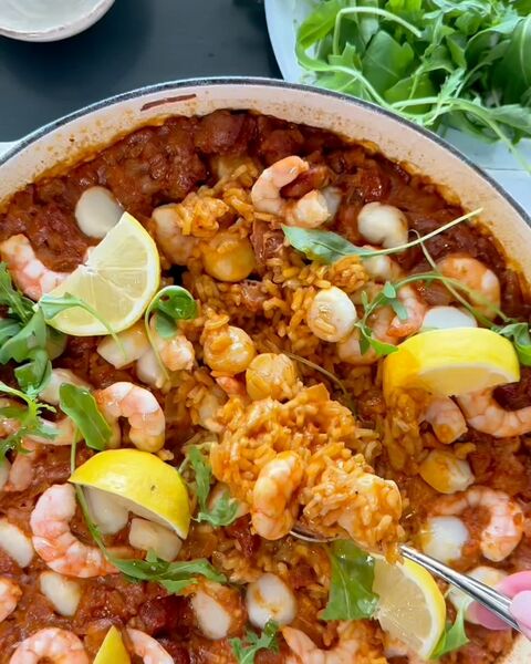 An overhead view of a white casserole of orange-red rice with prawns, scallops, lemon wedges and rocket, a spoonful lifted